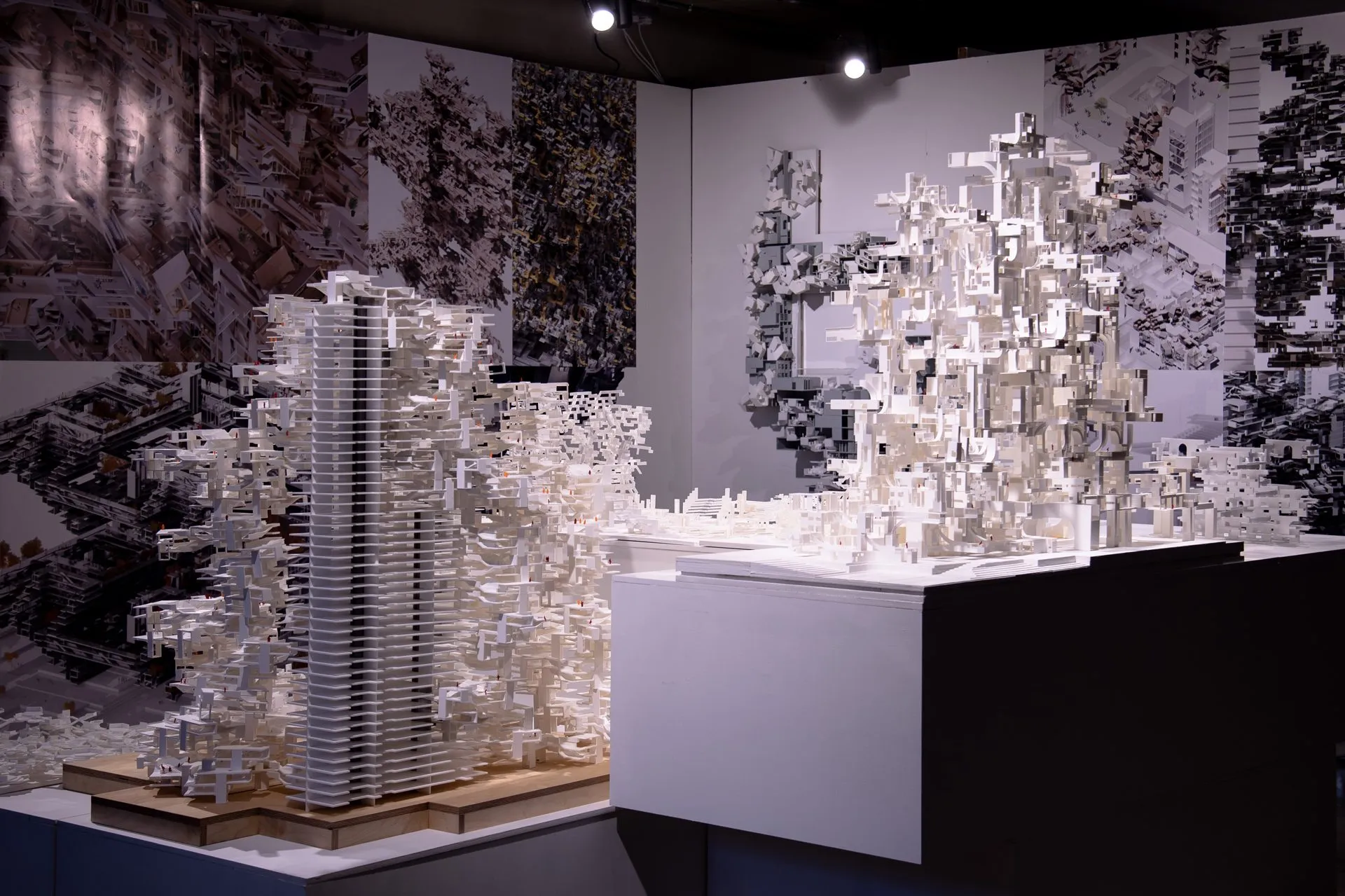 Two large white models, a tower of stacked slabs and a spreading aggregate, on plinths in front of wall-sized prints