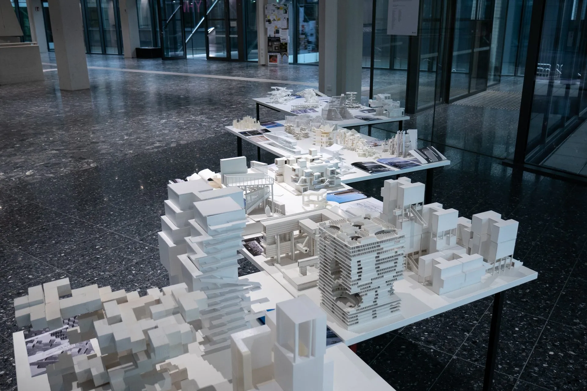 Tables of white models in the glazed atrium, with printed booklets laid between them