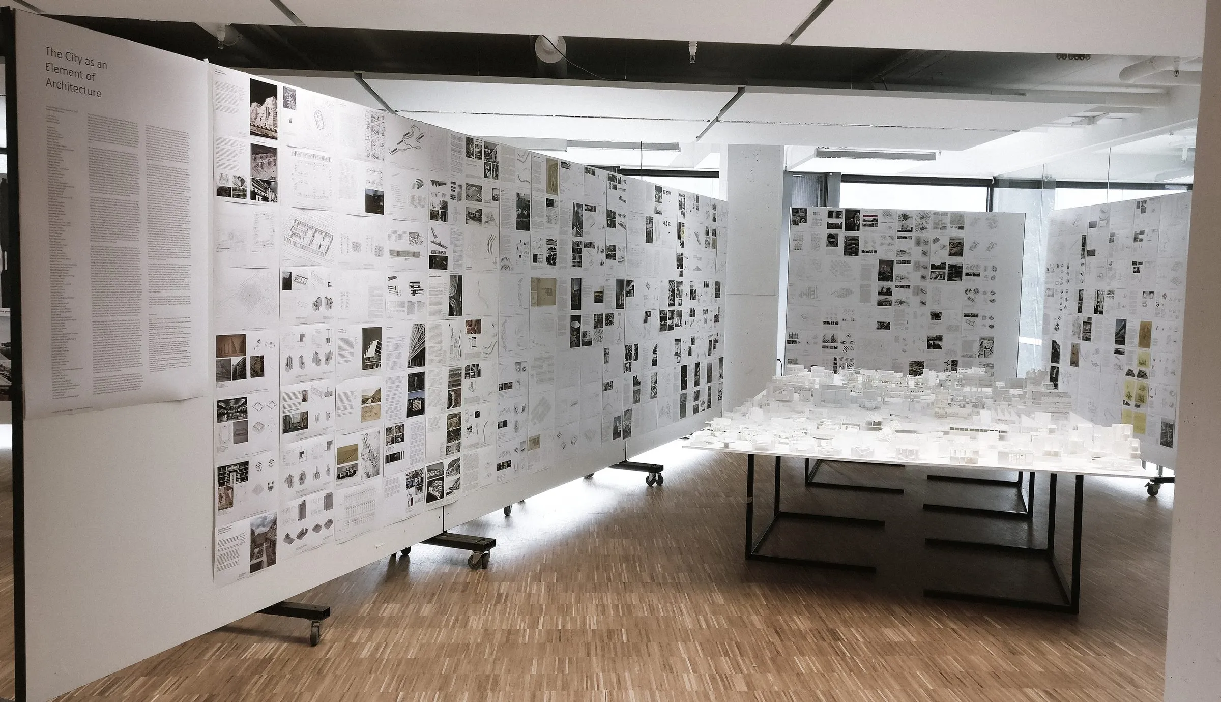 A long panel wall of pinned drawings running through the room, the table of white models behind it