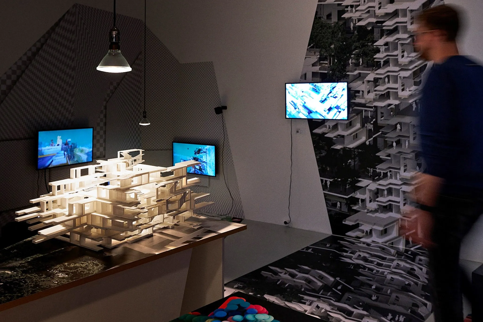 A lit white model of stacked housing floors on a plinth in a dark room, with screens and a wall print of the project behind it and a visitor passing