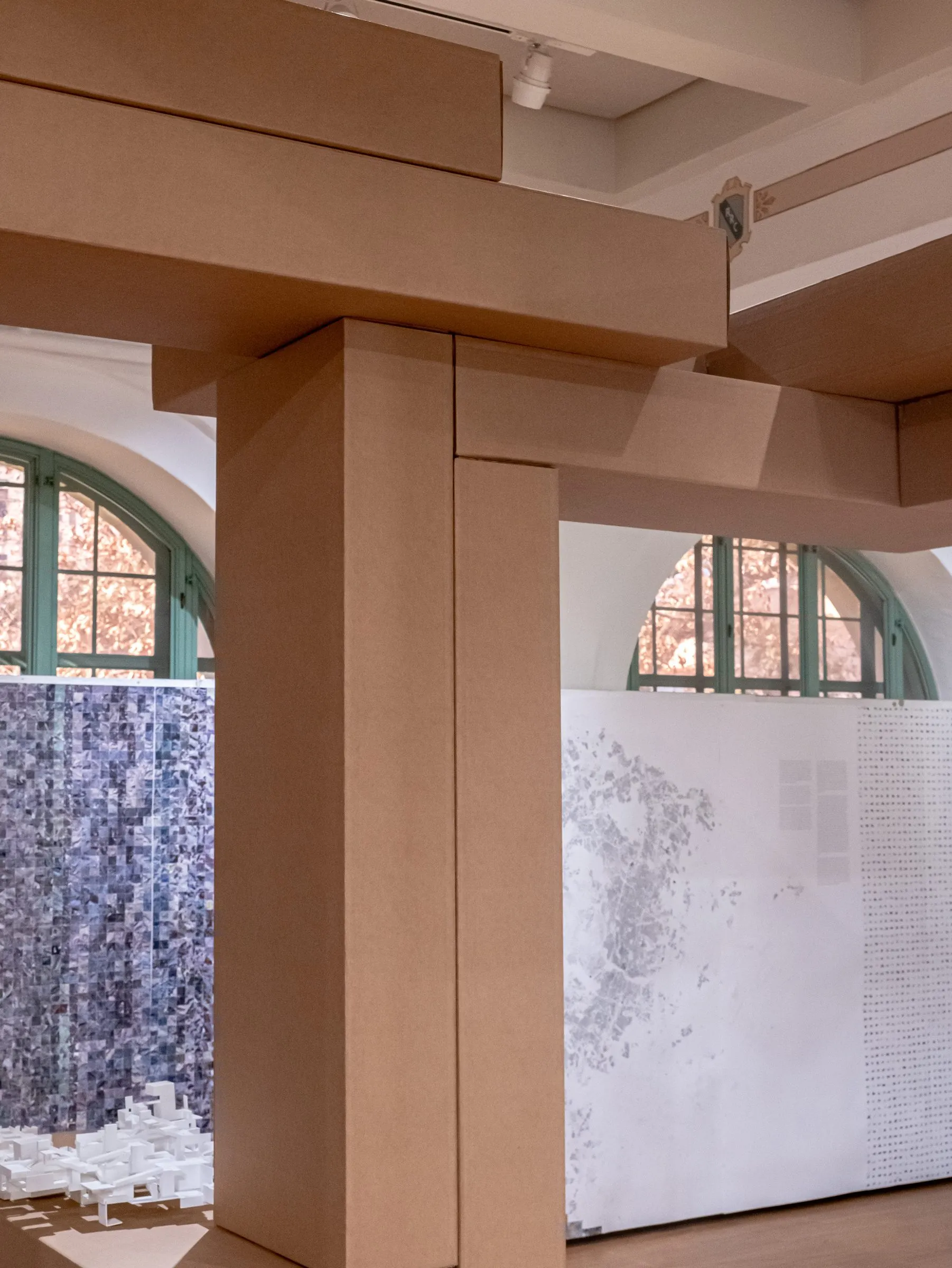 A cardboard column and beam before a printed wall, white models at its foot and an arched window at the side