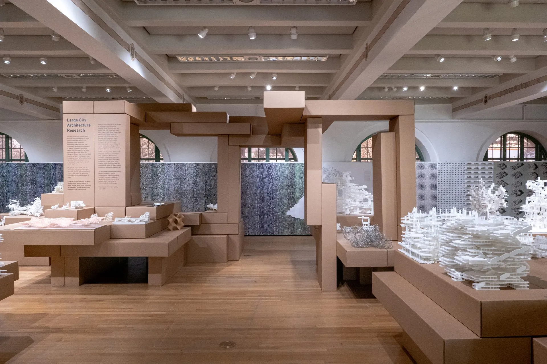 Wide view of the gallery: a cardboard portal frames tables of white models under a coffered ceiling