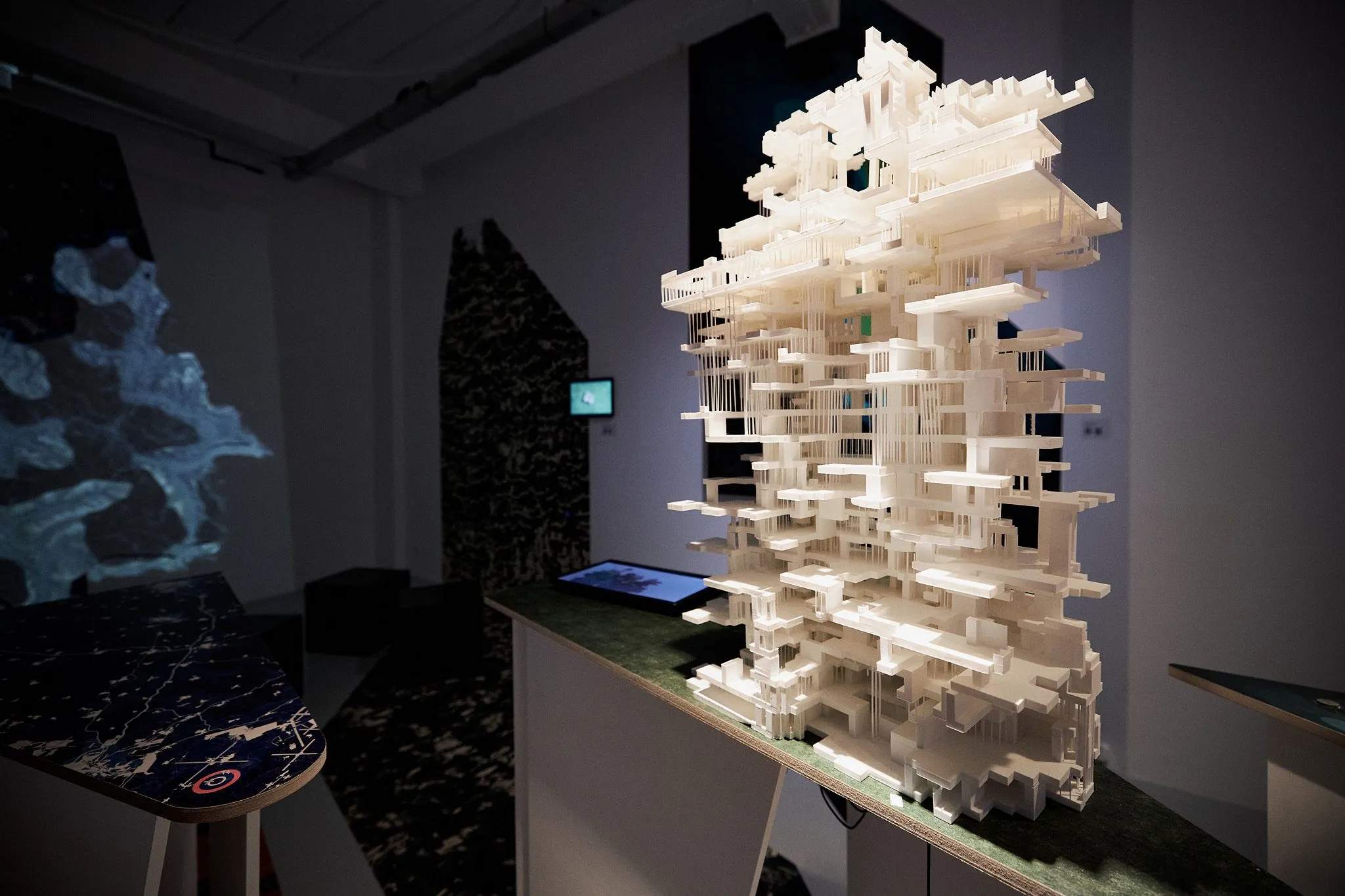 A white model of stacked slabs on a green-topped table, a screen on the table and a blue projection on the wall beside it