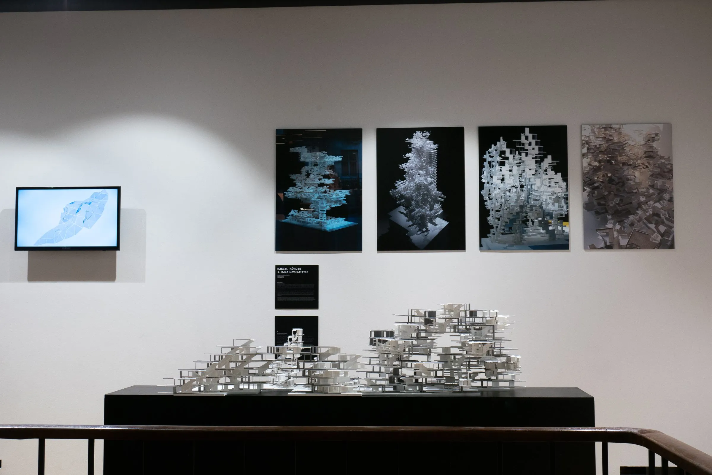 A gallery wall with a screen, four framed prints of the aggregates and a text panel; below it the white model on a black plinth