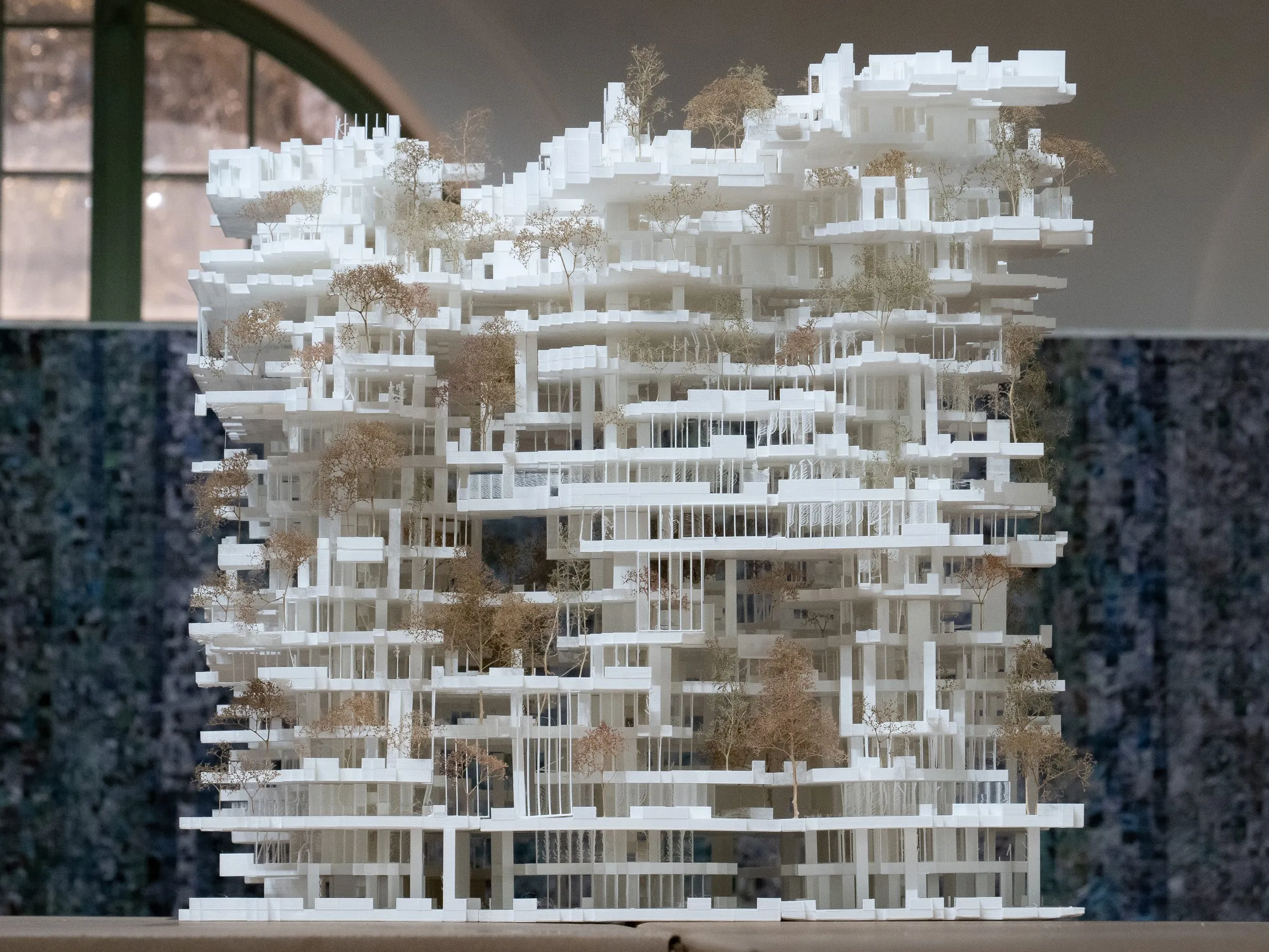 A white model of a tower of stacked slabs, columns and terraces with small dried trees, seen frontally in a gallery with arched windows