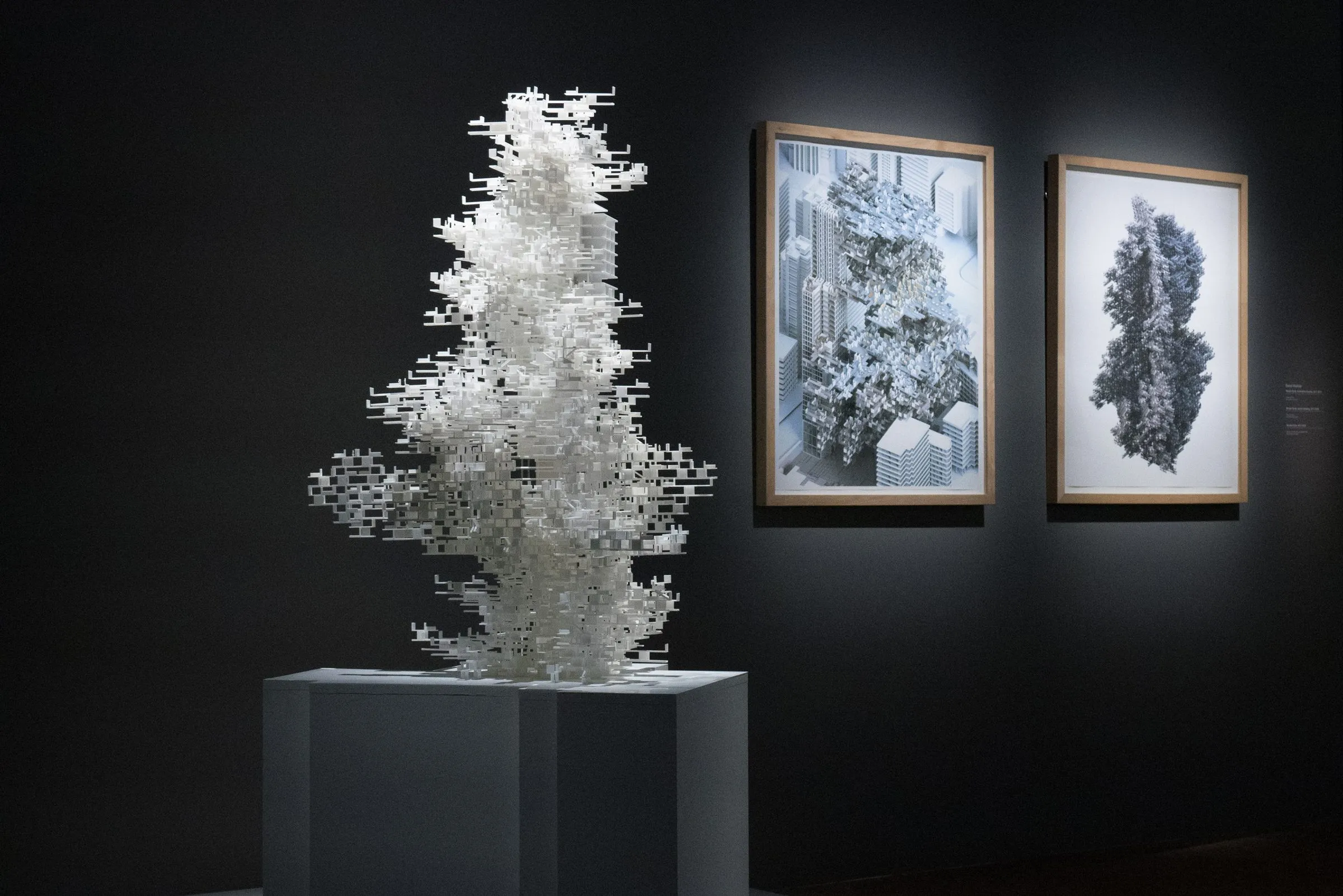 A white 3D-printed tower of courtyards on a pale plinth in a dark gallery, two framed drawings lit on the wall beside it